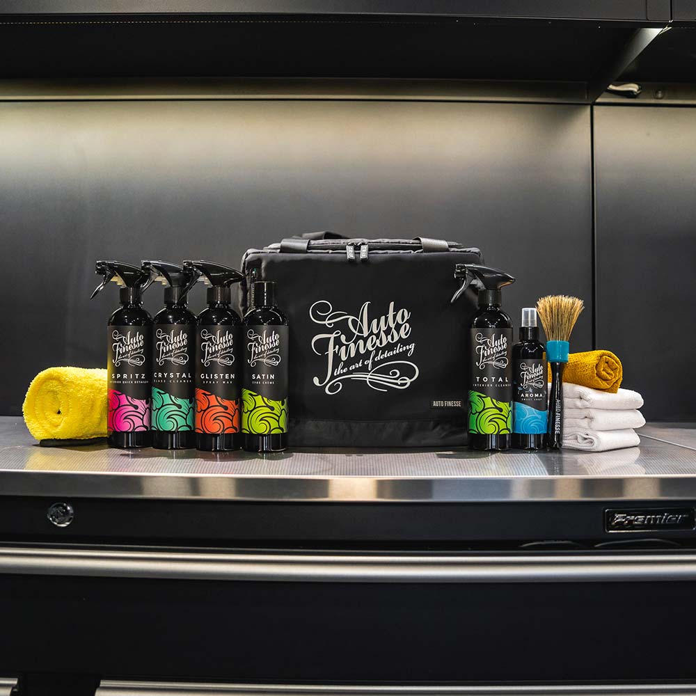 Automotive care products including bottles, a brush, and towels on a metallic surface with a branded bag.