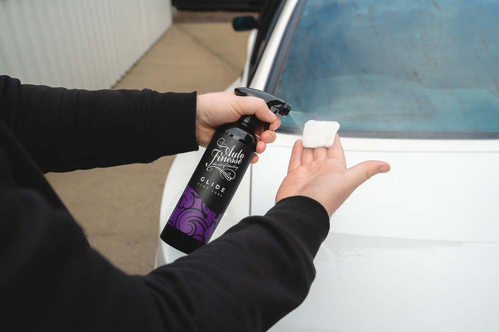 Person holding a car detailing spray bottle and a white applicator pad next to a car.