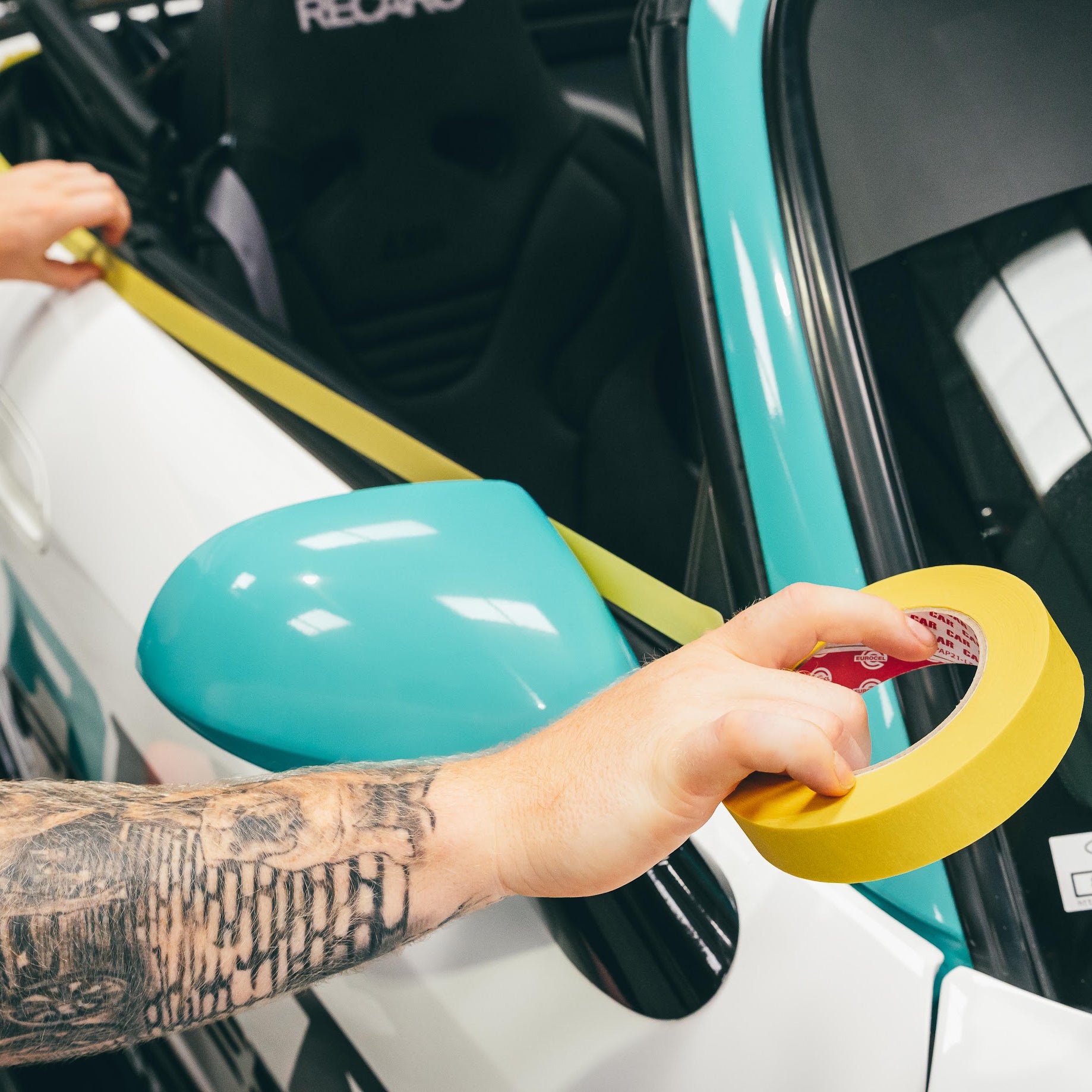 Person applying yellow tape to a car's side mirror with a visible tattoo on their arm.