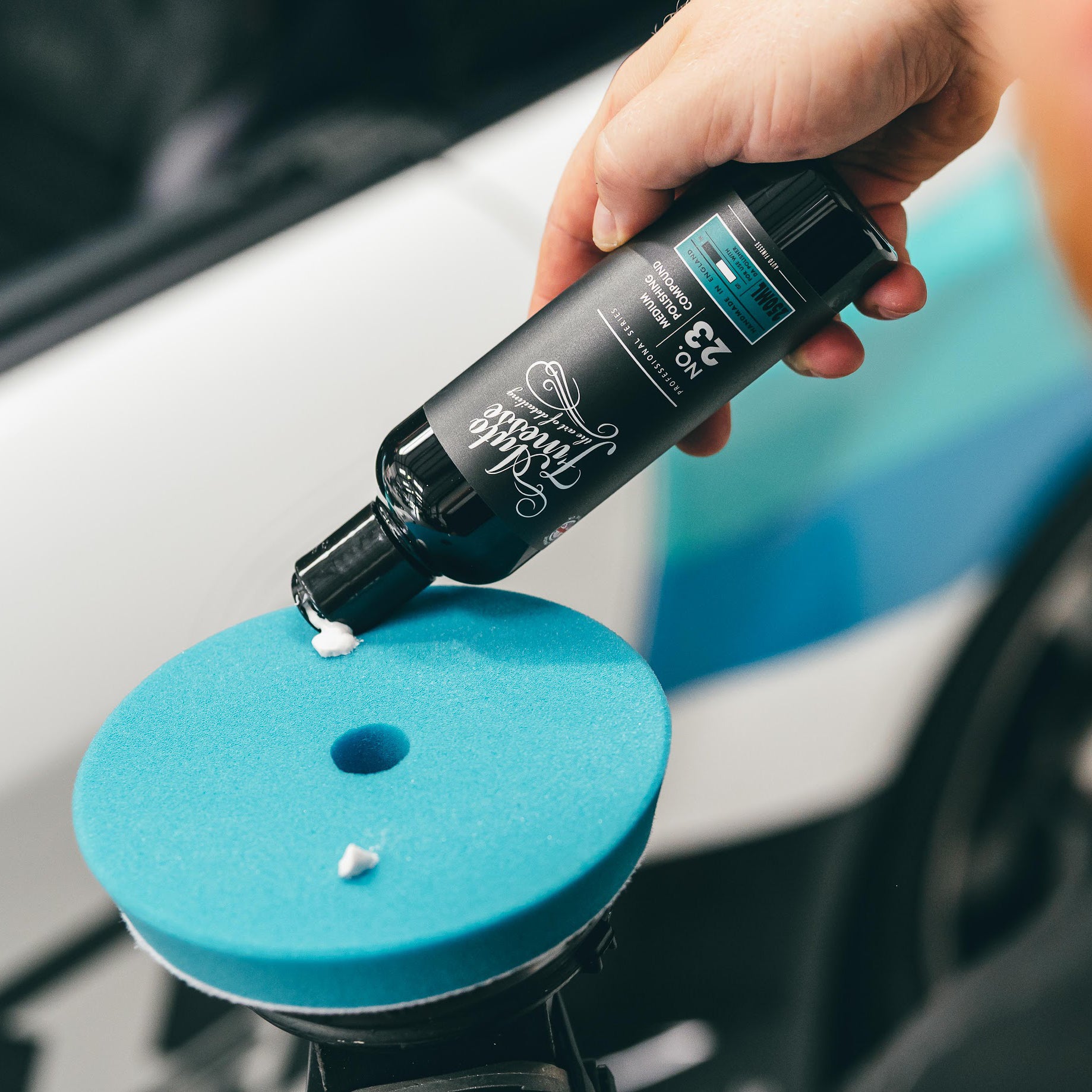 Hand holding a bottle of car care product over a blue foam pad on a car surface.